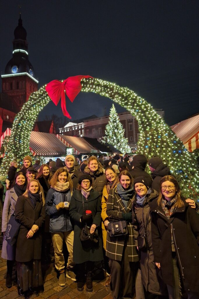 LJZA biedri sastājušies uz kopbildi pie Ziemassvētku tirdziņa vārtiem. Fonā redzams Doma baznīcas tornis un Ziemassvētku egle un lampiņas