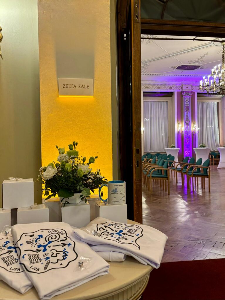 Latvijas Jauno zinātieku apvienības suvenīri - krūzītes, t-krekli ar saukli "Domā lēni!" un logotipa piespraudīes - dekoratīvi izvietotas uz galdiņa pie Rīgas Latviešu biedrības nama Zelta zāles ieejas.