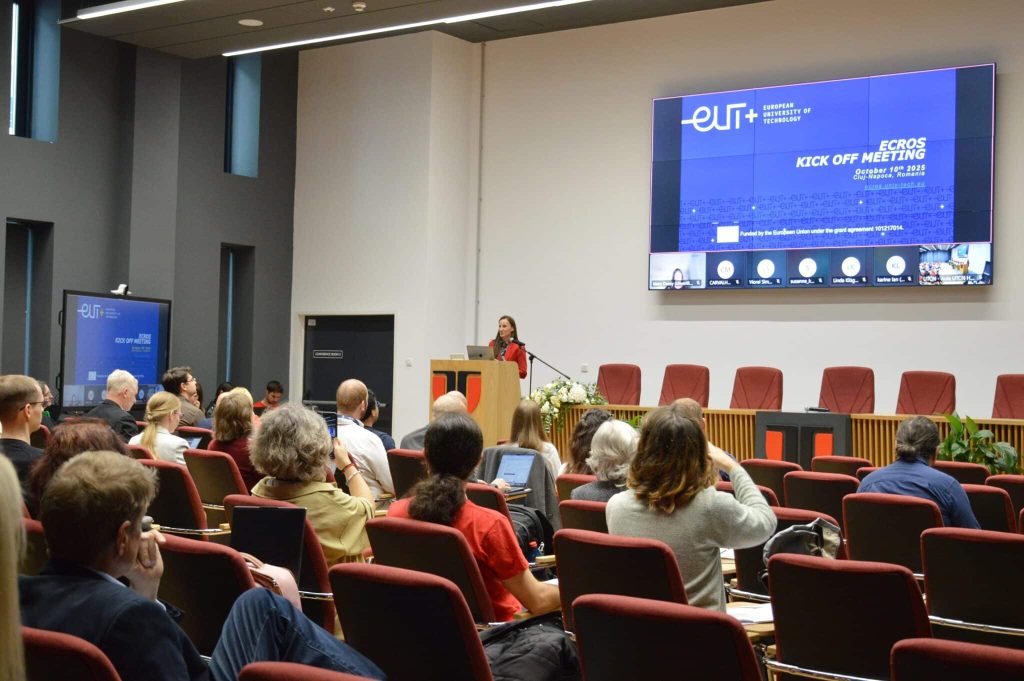 DSC_0597 Konferenču zālē sēž cilvēki un skatās prezentāciju. Auditorijas priekšā sieviete rāda slaidu ar ECROS projekta informāciju.